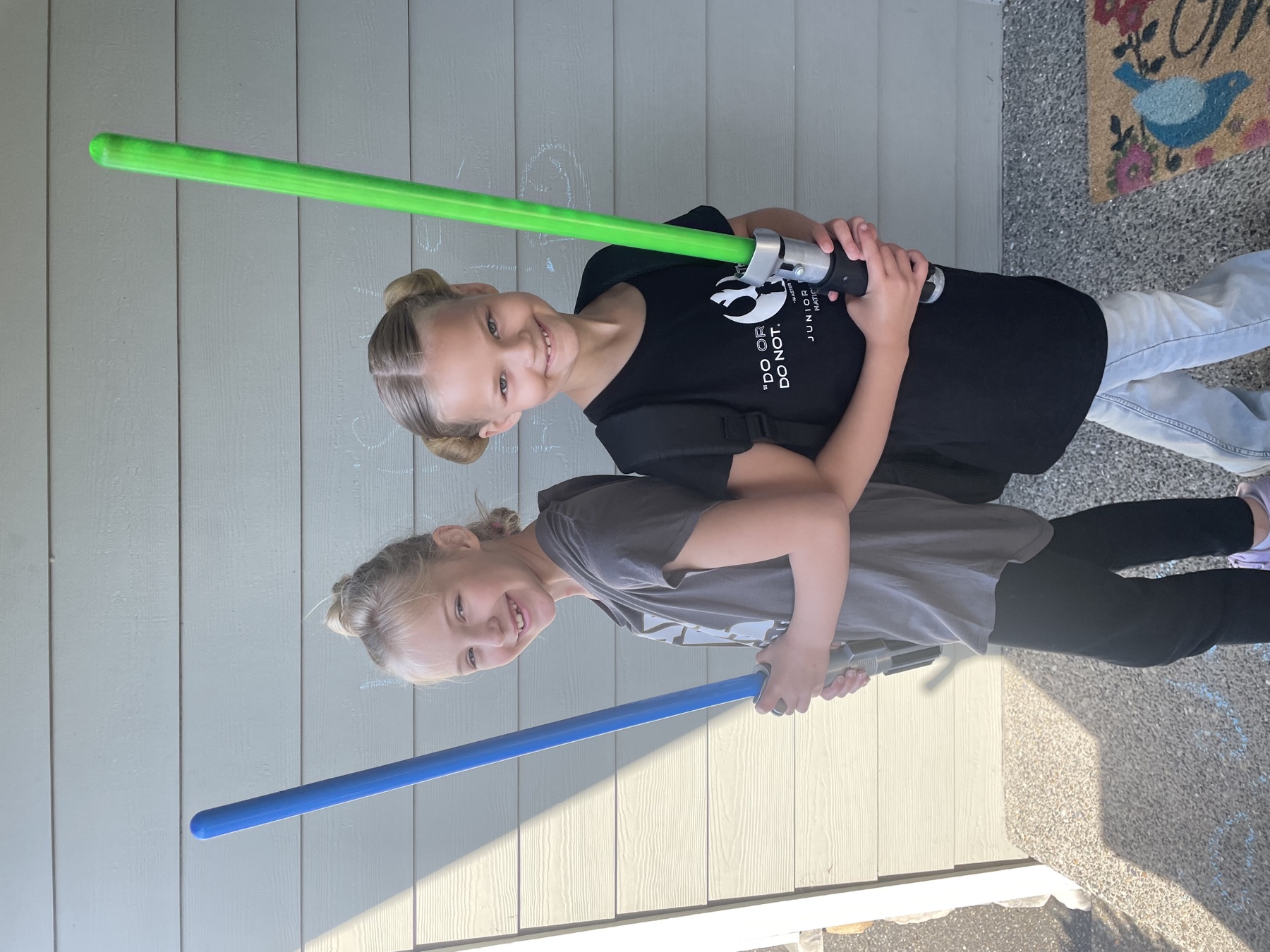 Hazel and her twin sister Ivy standing back to back, each holding a toy lightsaber, grinning