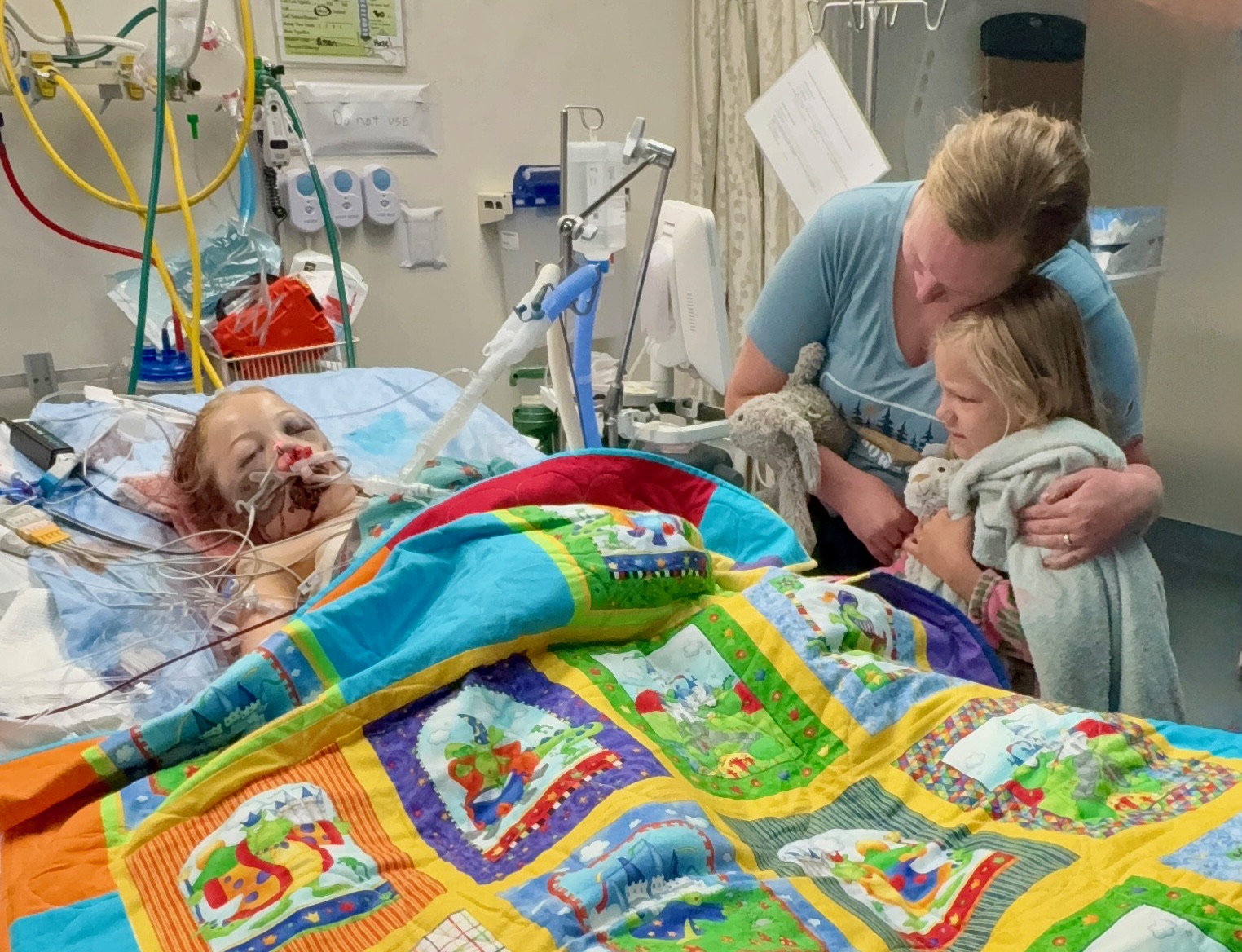 Hazel resting in her hospital bed at Harborview, with her bunny Hop Hop nearby, as her mom holds her sister close at the bedside